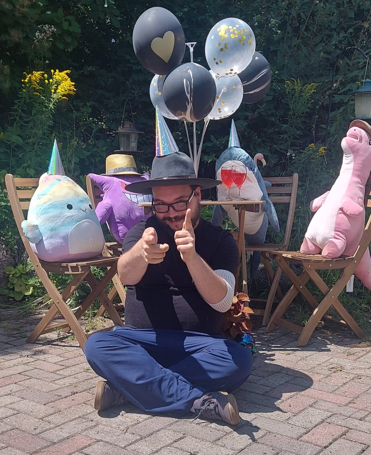 A man with facial hair and a felt sun hat sits on the ground in front of a patio set hosting a stuffed animal patio party, Photo 5